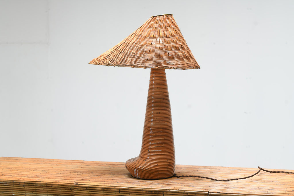 Chloe rattan table lamp with an asymmetric woven shade and tapered wrapped base on a rattan cabinet, with its power cord visible.