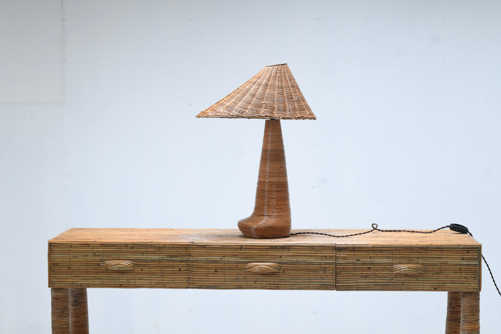 Chloe rattan table lamp with an asymmetric woven shade and curved wrapped base on a three-drawer rattan console, with its cord and inline switch visible.