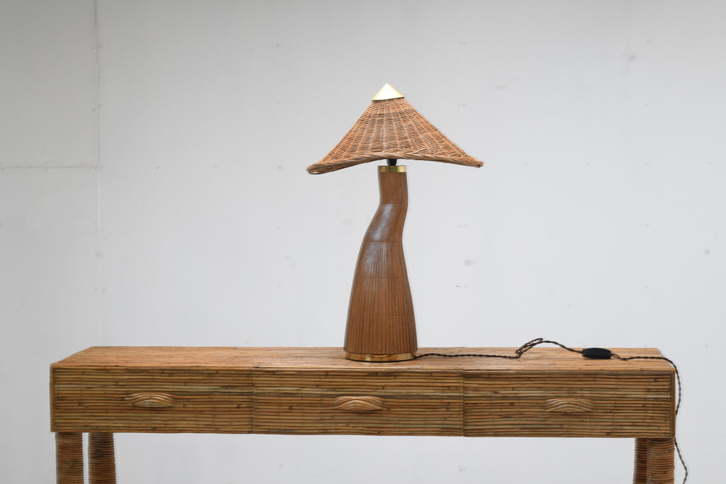 Shroom rattan table lamp with a woven peaked shade and curved ribbed base on a three-drawer rattan console, with its cord and inline switch visible.