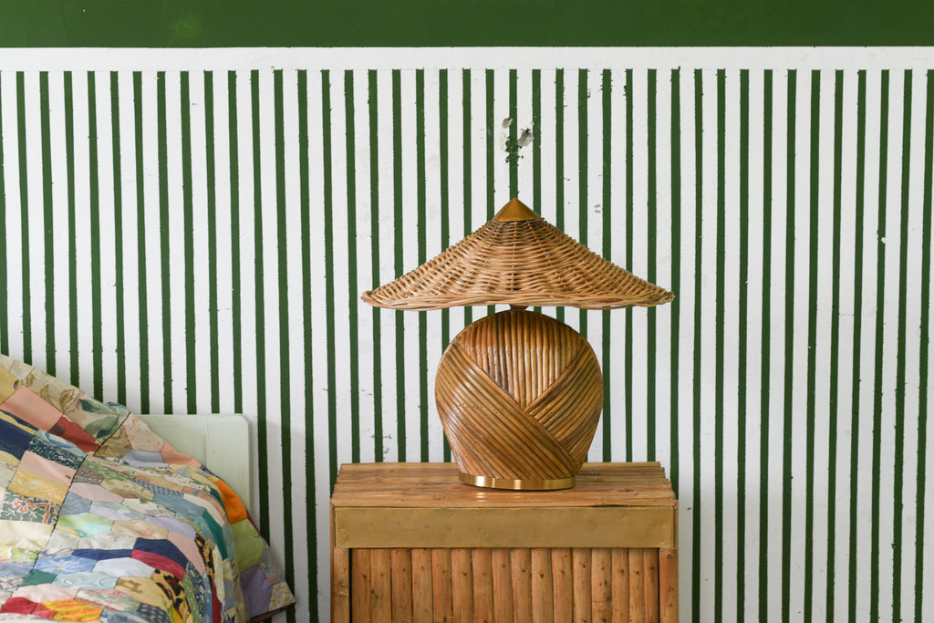 Deka rattan table lamp on a wooden bedside cabinet beside a bed, against a green-and-white striped wall.