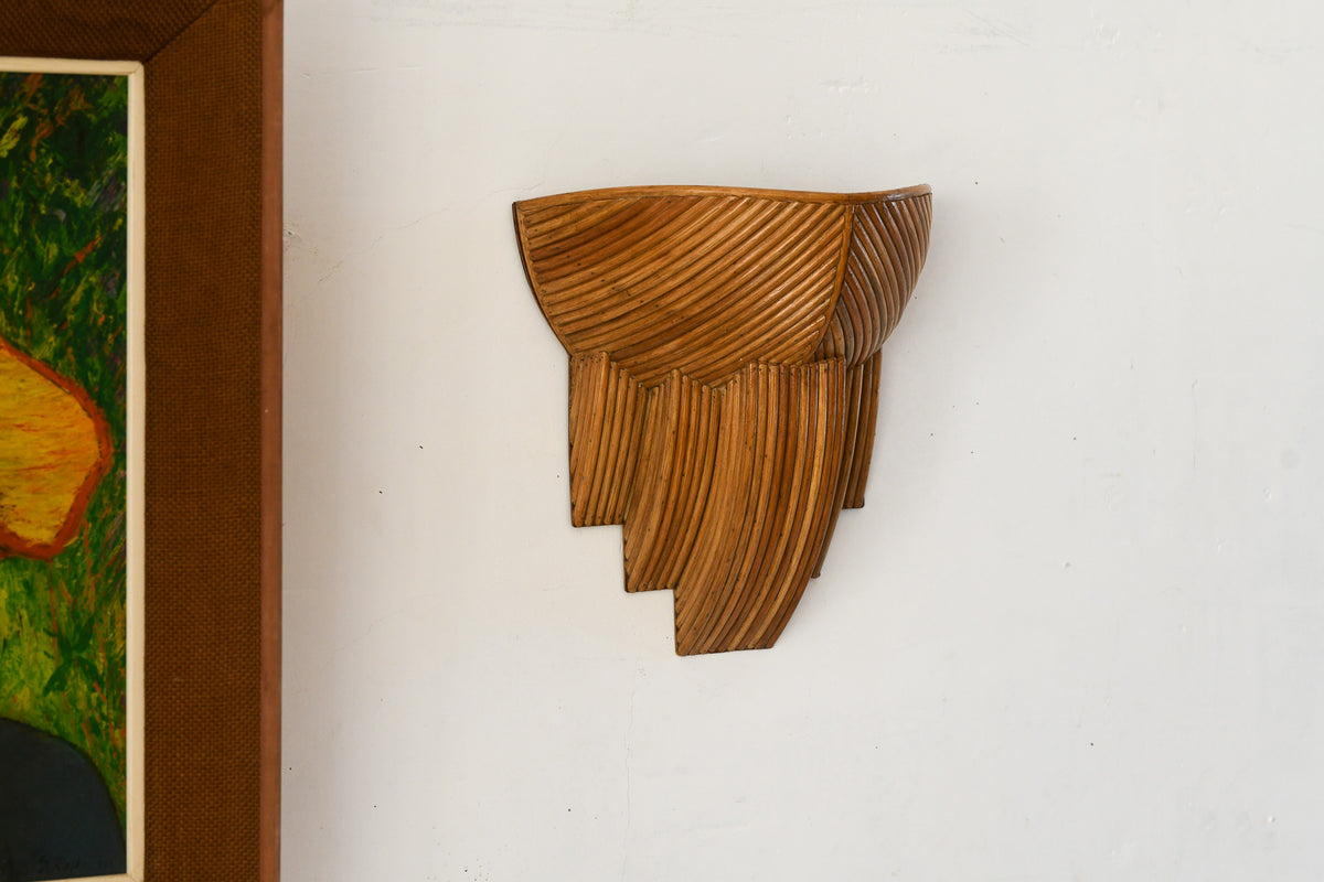 Barbara rattan wall sconce with diagonal reed detailing and stepped lower sections, mounted on a white wall beside framed artwork.